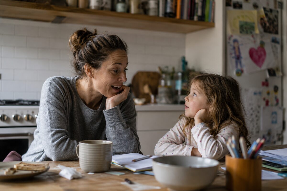 A mother and young daughter talking at the kitchen table with a notebook open.